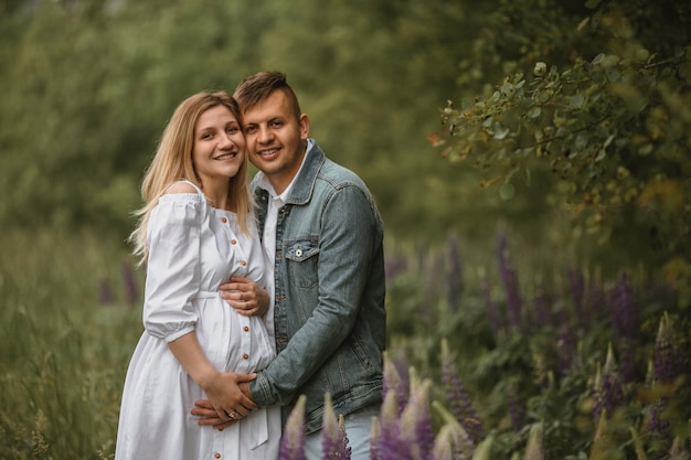 Vooraanzicht van tedere toekomstige ouders die knuffelen in een groene tuin met violette lupines, er recht en glimlachend uitziend