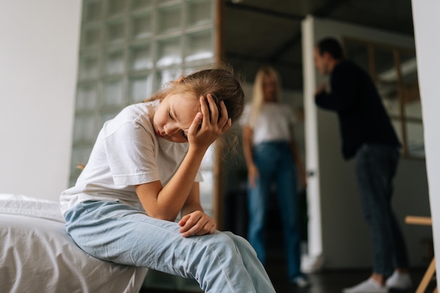 Vooraanzicht van verdrietig eenzaam meisje wegkijkend huilend lijden zittend op de bank terwijl ouders ruzie maken en vechten in de woonkamer op de achtergrond