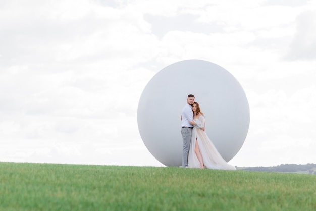 Vooraanzicht van verliefde pasgetrouwden staan op de achtergrond van een wit monument in de vorm van een bal in het midden van het veld