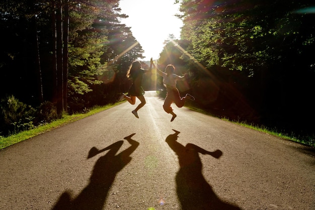 Vooraanzicht van vrienden die midden op de weg springen. horizontale weergave van vrouwen die in tegenlichtschaduwen springen die op de achtergrond van de natuur reizen. zomer vakantie concept.