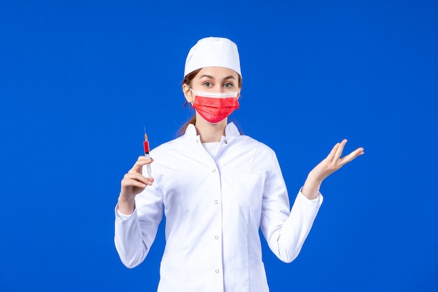 Vooraanzicht vrouwelijke verpleegster in wit medisch pak met rood masker en injectie in haar handen op blauw