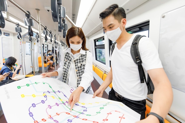 Vooraanzichtportret van jonge volwassen Aziatische paartoeristen met een medisch masker die staan en de papieren metrokaart bij elkaar houden in de Skytrain met een wazige kaartvoorgrond en Skytrain-achtergrond