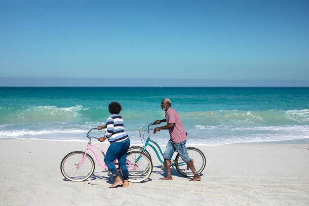 Voorbeeld van een oud Afro-Amerikaans echtpaar dat op het strand staat met een blauwe hemel op de achtergrond, op hun fietsen rijdt, praat en glimlacht naar elkaar.