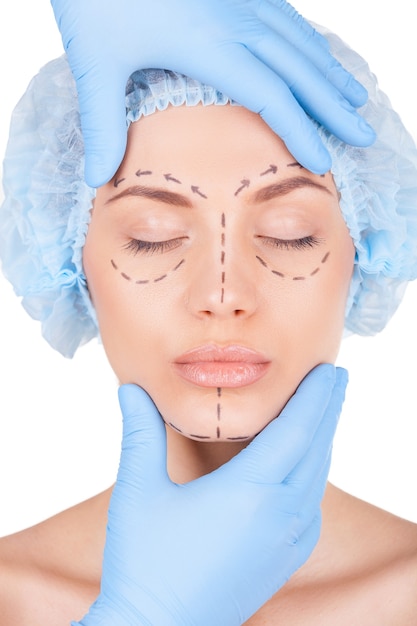 Voorbereiding voor gezichtschirurgie. Aantrekkelijke jonge vrouw met medische hoofddeksels en schetsen op het gezicht die de ogen gesloten houden terwijl de arts haar gezicht onderzoekt dat op wit wordt geïsoleerd