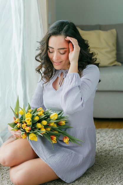 Voorjaarsportret van mooie donkerharige gekrulde vrouw in lila jurk met groot kleurenboeket tulpen thuis Emotioneel model houdt bloemen in haar handen Vrouwendag Aanwezig Moederdag