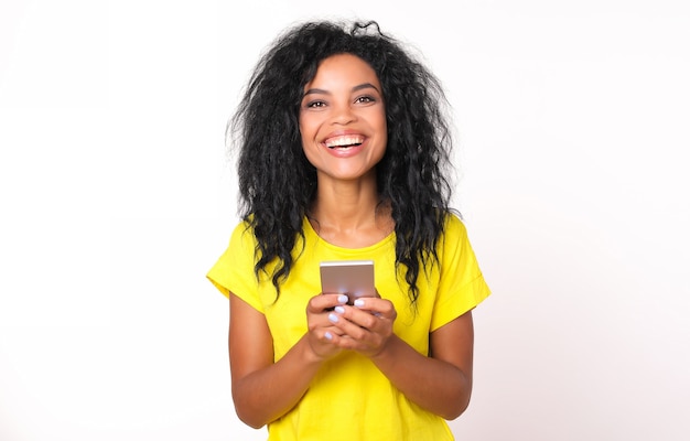 Voorportret van een vrolijk Afro-Amerikaans meisje met schouderlang rommelig zwart haar, dat oprecht lacht terwijl ze een smartphone in haar handen houdt