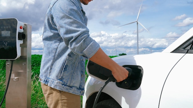 Vooruitstrevende man met zijn elektrische auto Ev-auto die energie oplaadt van laadstation op groen veld met windturbine als concept van toekomstige duurzame energie Elektrisch voertuig met energiegenerator