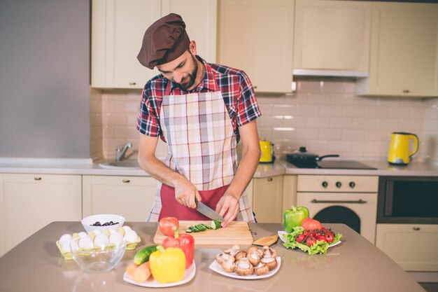 Voorzichtig koken staat en snijd komkommer in stukjes. Hij doet het met een mes. Guy kijkt naar beneden. Hij is serieus en geconcentreerd.