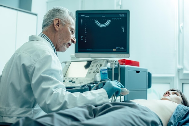 Voorzichtige mannelijke echografist zit naast de jonge man op een medische bank en kijkt naar hem terwijl hij een ultrasone transducer vasthoudt