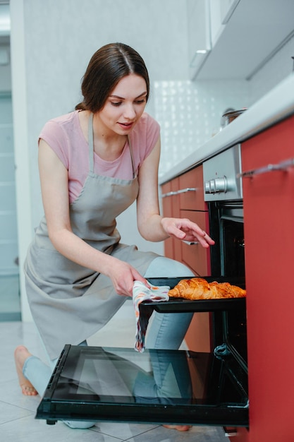 Voorzichtige vrouw in een schort die naar de open oven leunt en naar de croissants op een bakpan kijkt