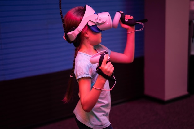 Виртуальная игра и виртуальная реальность, девочка-геймер восьми лет, весело играющая в футуристические видеоигры-симуляторы в 3D-очках и джойстиках в развлекательной виртуальной комнате, инновационные технологии и неоновый свет