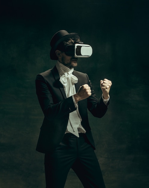 VR spelen. Jonge man in pak als Dorian Gray geïsoleerd op donkergroene achtergrond. Retro stijl, vergelijking van tijdperken concept. Mooi mannelijk model als klassiek literatuurkarakter, ouderwets.