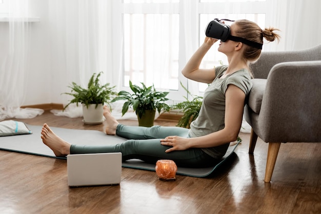 Фото Молодая женщина сидит в положении лотоса в наушниках виртуальной реальности, занимаясь йогой.