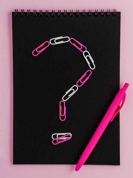 Vraagteken gemaakt van clips op zwarte notebook leeg op roze achtergrond.