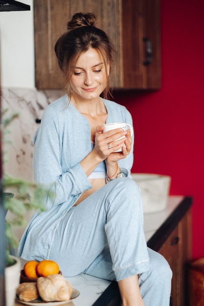 Vreedzame brunette vrouw in pyjama's met een kopje koffie en zittend op het aanrecht.