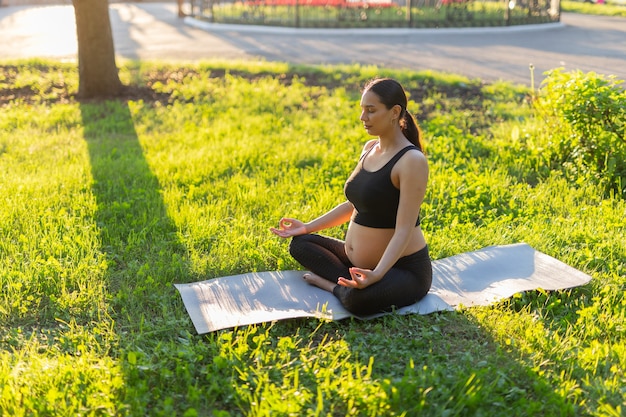 Vreedzame jonge positieve zwangere vrouw in gymnastiek pak doet yoga en mediteren zittend op de mat op groen gras op zonnige warme zomerdag. concept voorbereiding op de bevalling en een positieve houding