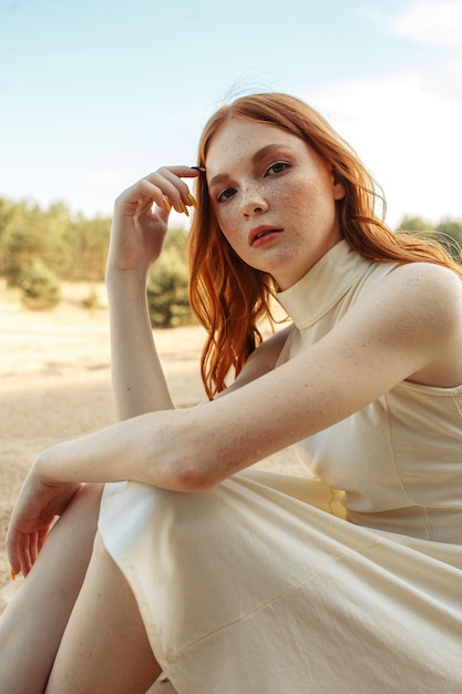 Vreedzame jonge vrouw met rood haar in zomerjurk zittend op het zandstrand en kijkend naar de camera