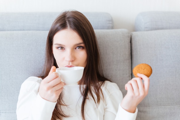 Vreedzame mooie jonge vrouw zittend op een grijze bank en thee drinken met cookie