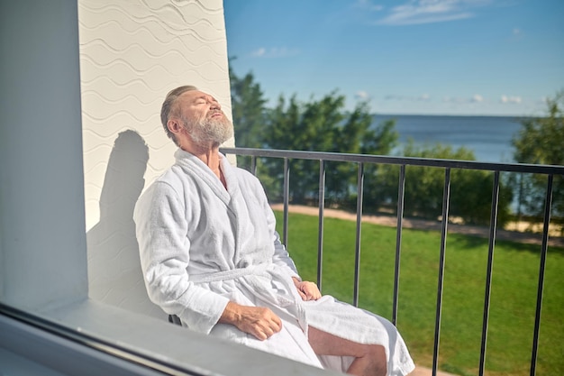 Vreedzame vakantieganger in de witte badstof badjas zonnebadend zittend op de stoel op het balkon