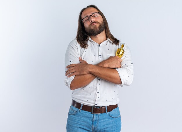 Vreedzame volwassen knappe man met een bril die een winnaarbeker vasthoudt en de handen gekruist houdt met gesloten ogen geïsoleerd op een witte muur met kopieerruimte