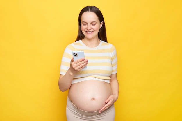 Foto vreemde blanke zwangere vrouw met blote buik die een casual top draagt geïsoleerd over een gele achtergrond haar buik streelt en met behulp van een mobiele telefoon berichten leest op een forum voor zwangere vrouwen