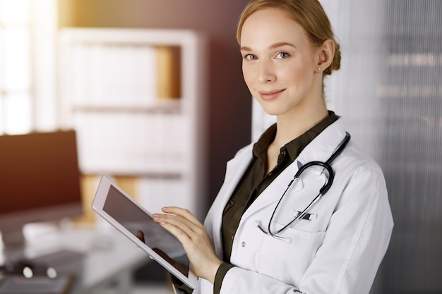 Vreemde glimlachende vrouwelijke arts met een tabletcomputer in een zonnige kliniek. Portret van een vriendelijke artsvrouw op het werk. Perfecte medische dienst in het ziekenhuis. Medicijnconcept.