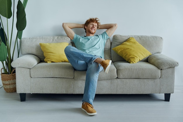Vreemde roodharige man die zijn handen achter zijn hoofd houdt terwijl hij thuis op de bank ontspant