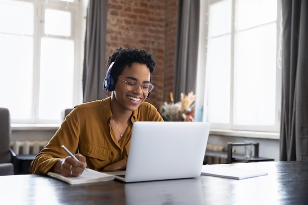Vreemde zwarte universiteitsstudent met een koptelefoon die een virtuele les bijwoont, aantekeningen schrijft, een webinar op internet kijkt, online studeert, Afrikaanse werknemer met een hoofdtelefoon die met een klant praat via een videogesprek