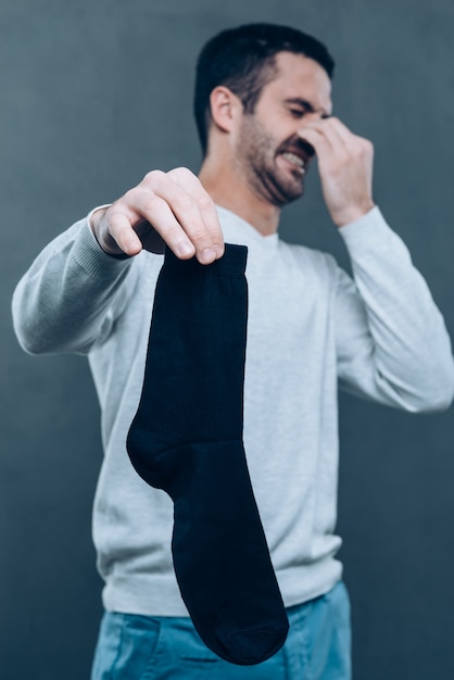 Vreselijke geur! Gefrustreerde jongeman die negativiteit uitdrukt en zijn neus bedekt met vingers terwijl hij een zwarte sok vasthoudt en tegen een grijze achtergrond staat