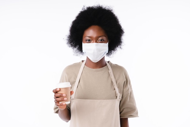 Vreugdevol Afrikaans meisje barista in medisch masker tegen Covid-19 met een kop koffie geïsoleerd in het wit