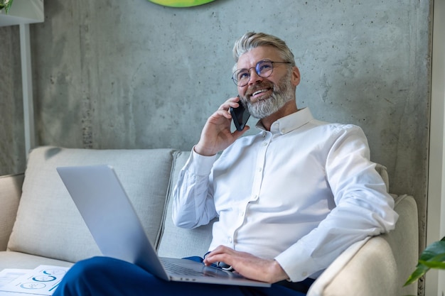 Foto vreugdevol volwassen zakenman met de draagbare computer zit op de bank tijdens het telefoongesprek