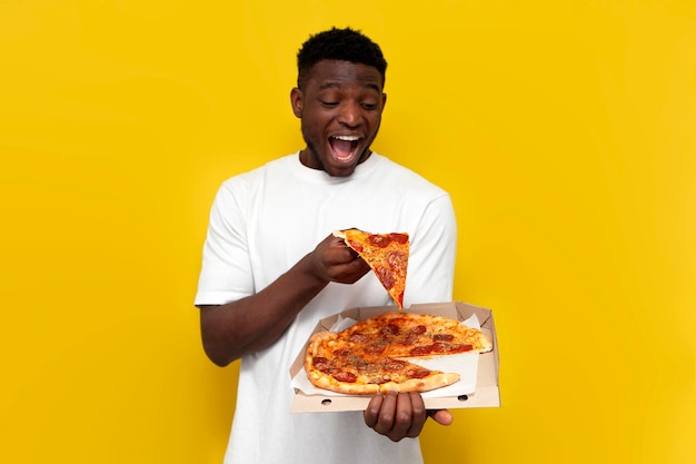 Vreugdevolle Afrikaans-Amerikaanse man in wit T-shirt houdt een doos pizza vast op een gele geïsoleerde achtergrond
