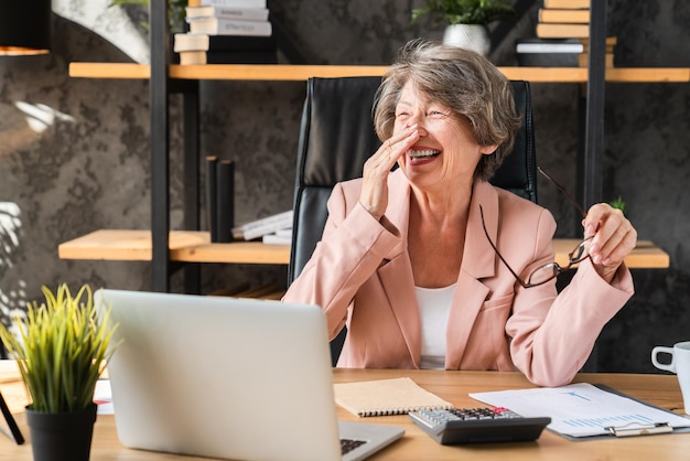 Vreugdevolle blanke senior zakenvrouw in formele kleding die lacht terwijl ze aan de laptop werkt
