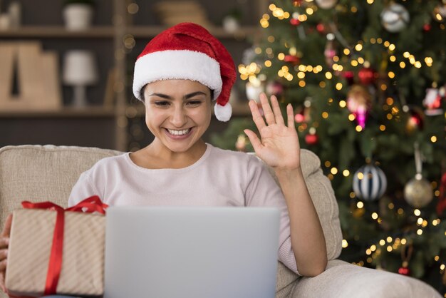 Vreugdevolle Indiase vrouw die met haar hand zwaait en van een webcam videogesprek op de computer geniet. Ze feliciteert familie of vrienden met het nieuwe jaar online en houdt een verpakt geschenkdoosje vast. Vrolijk kerstfeest.