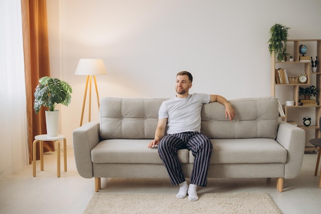 Vreugdevolle jonge baarde man in pyjama zit op een bank in de woonkamer thuis met kopieerruimte