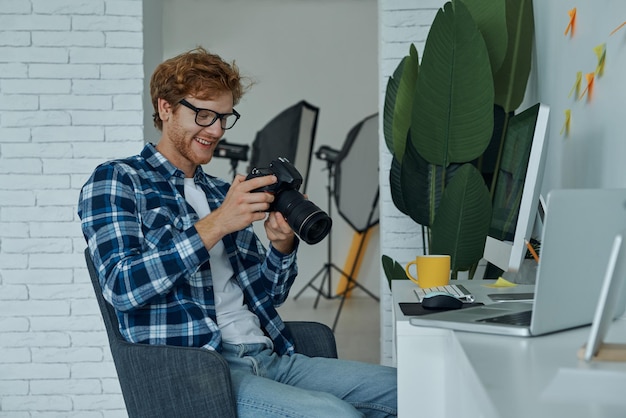 Vreugdevolle jonge man die de camera onderzoekt terwijl hij op zijn werkplaats in de studio zit