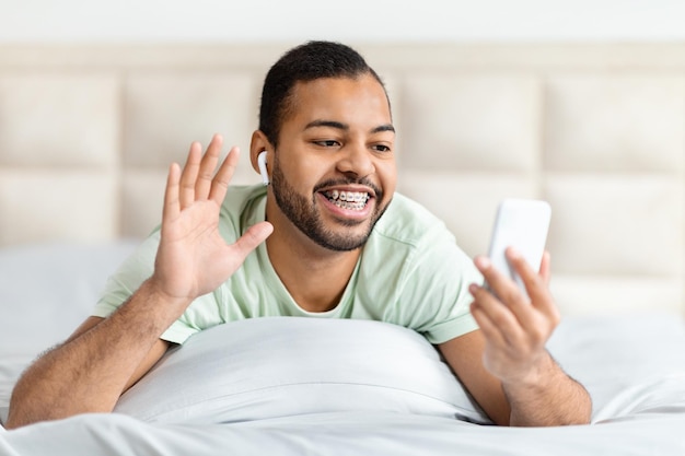 Vreugdevolle jonge zwarte man ligt op bed hebben videogesprek