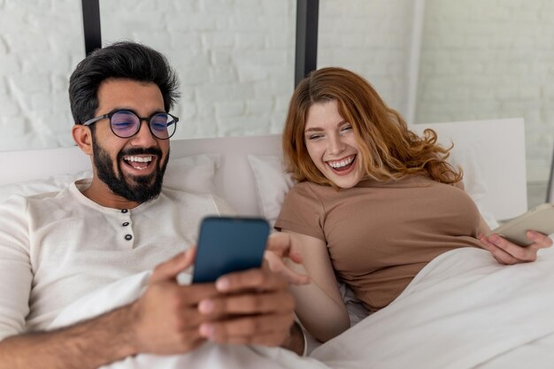 Foto vreugdevolle man en vrouw die het weekend samen in bed doorbrengen met hun mobiele telefoon en plezier maken met lachen