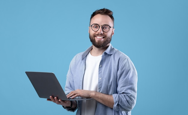 Vreugdevolle millennial man met een bril die een laptop gebruikt voor online werk of communicatie op blauw