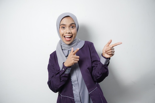 Vreugdevolle mooie Aziatische vrouw in casual shirt en hijab wijst naar een lege ruimte geïsoleerd door een witte achtergrond