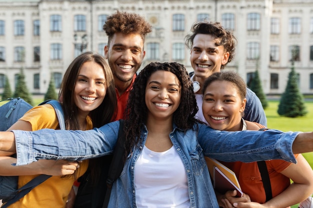 Vreugdevolle multiculturele studenten nemen een selfie bij het universiteitsgebouw en poseren samen in de open lucht.