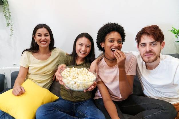 Vreugdevolle multiculturele vrienden die samen een film kijken, glimlachend popcorn delen op een gezellige bank.