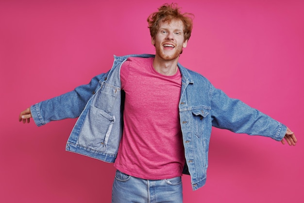 Vreugdevolle roodharige man in denim kleding die tegen een roze achtergrond staat