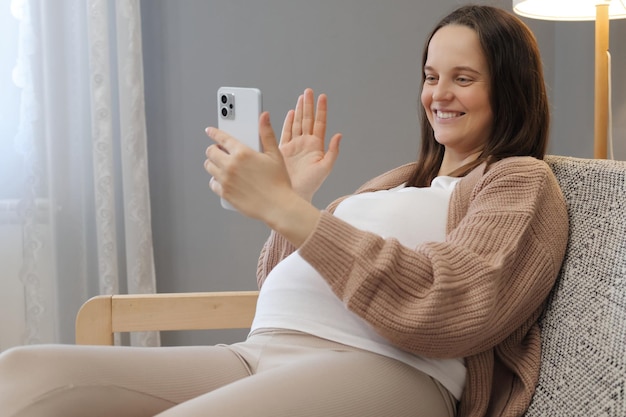 Vreugdevolle, verheugde zwangere vrouw zit op de bank met haar mobiele telefoon, houdt haar hand op haar buik, glimlacht, heeft een online videogesprek, zwaait met haar hand en toont een begroetingsgebaar.
