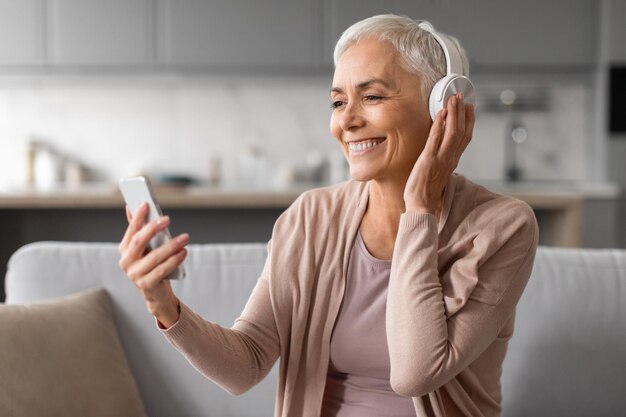 Vreugdevolle volwassen vrouw met mobiele telefoon met draadloze koptelefoon binnen