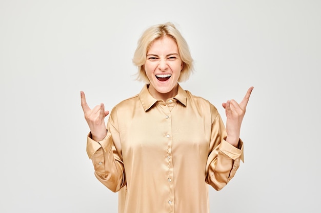 Vreugdevolle vrouw in een beige blouse die rock and roll handgebaar maakt geïsoleerd op een witte achtergrond