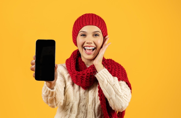 Vreugdevolle vrouw in winter breiwerk toont telefoon lege scherm studio