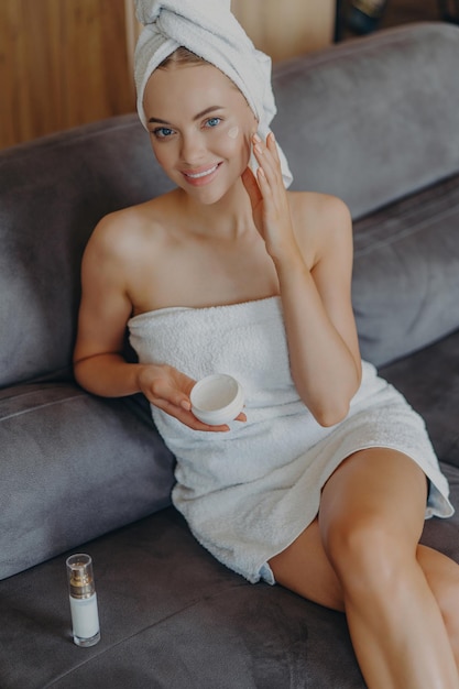 Vreugdevolle vrouw past gezichtscrème aan en glimlacht gewikkeld in een handdoek op de bank thuis