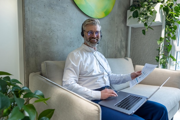 Vreugdevolle zakenman in de headset zit op de bank met documenten in de hand die op zijn laptop tikken