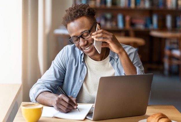 Vreugdevolle zwarte man die op zijn smartphone praat en online werkt in een café.